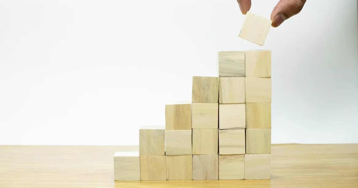 A hand places a wooden block on top of a rising staircase of blocks